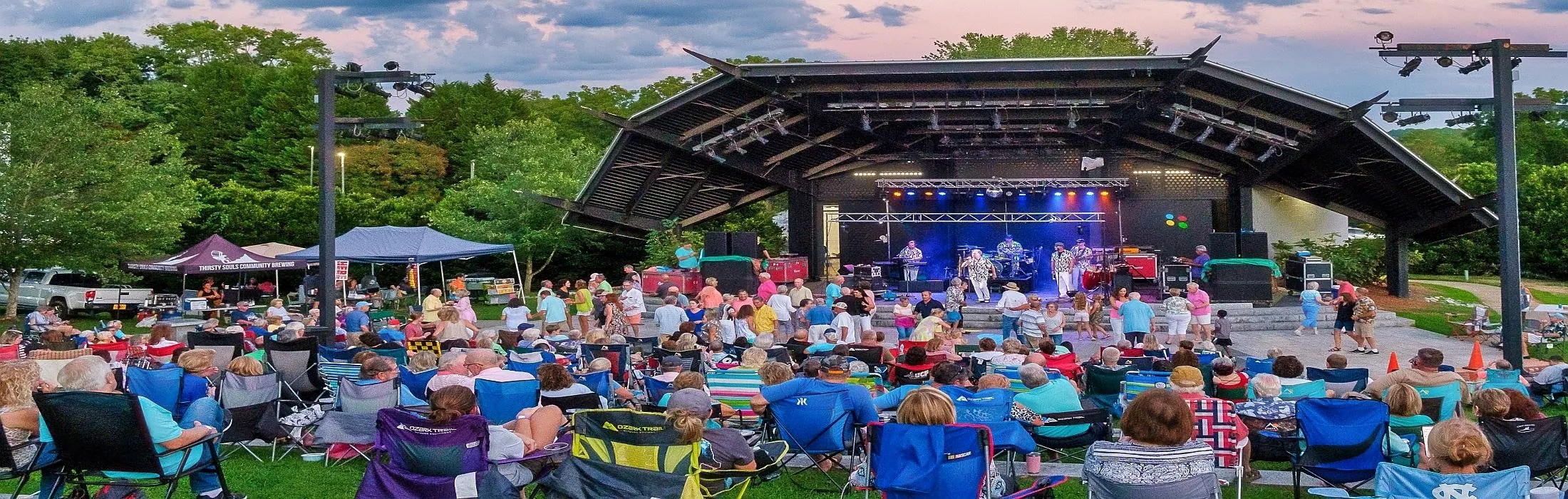 A crowd in lawn chairs enjoys a live outdoor concert at the Mount Airy amphitheater at dusk