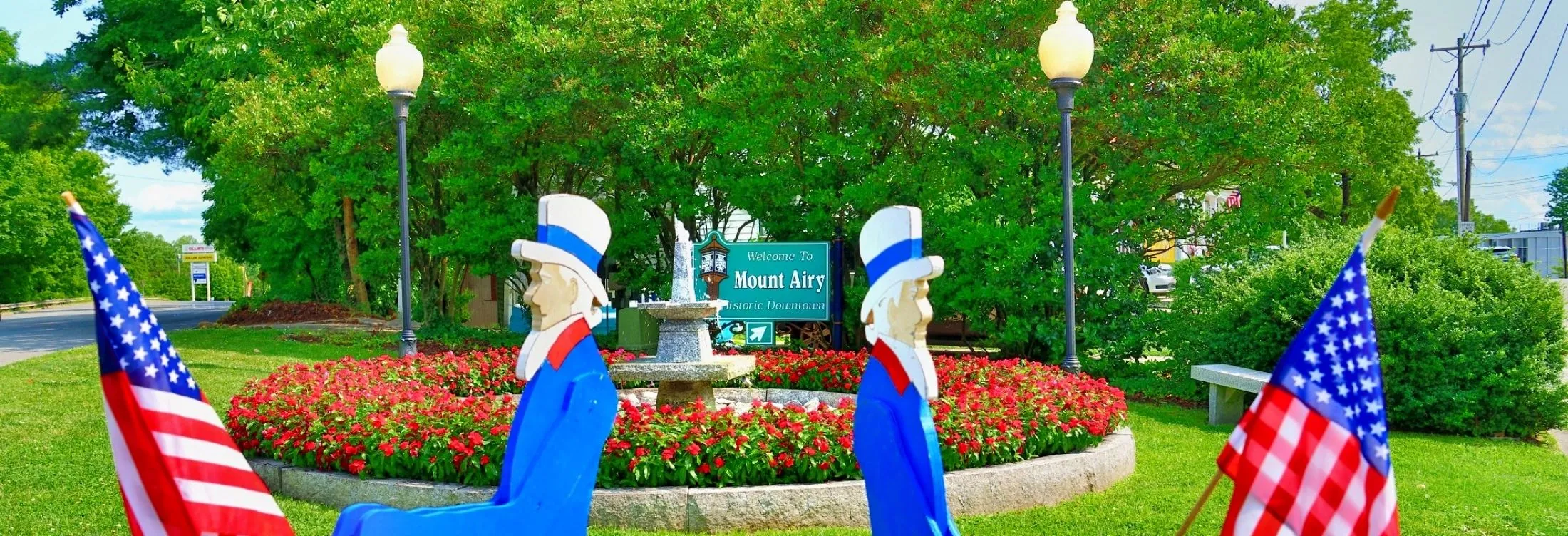 Welcome to Mount Airy sign and fountain in a downtown park, framed by American flags and summer flowers
