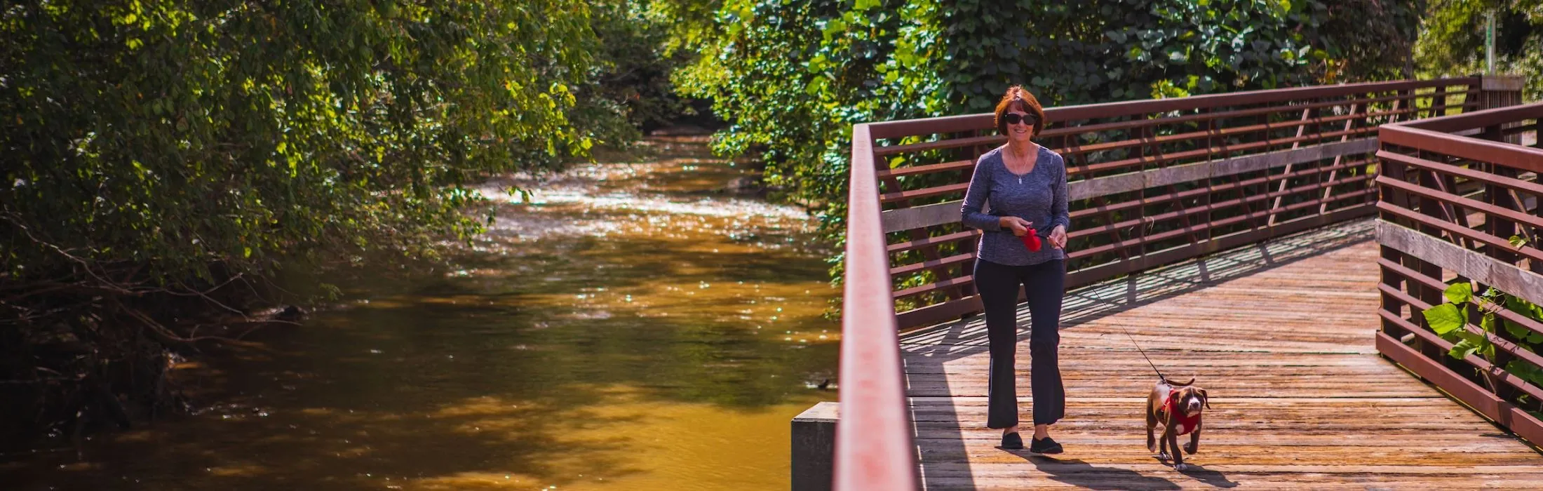 A resident walks her dog across a wooden bridge over the river on the Granite City Greenway